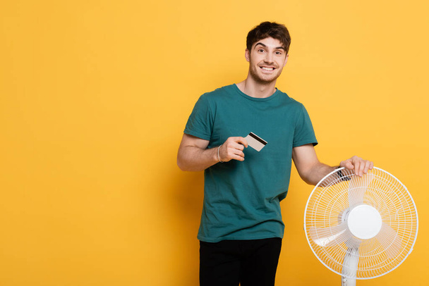 happy man holding credit card while standing with electric fan on yellow      - Fotoğraf, Görsel