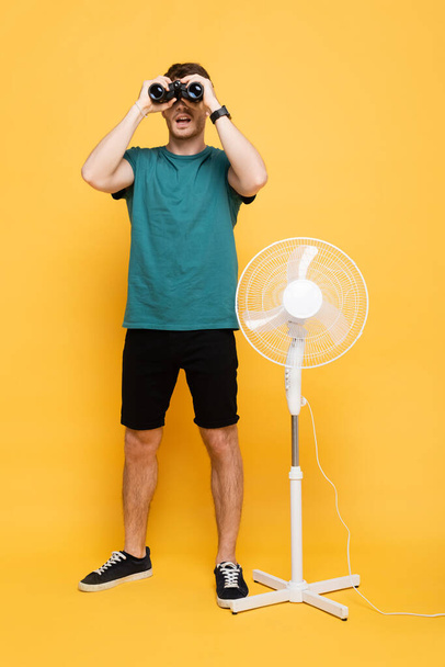 young man looking through binoculars while standing with electric fan on yellow      - Photo, Image