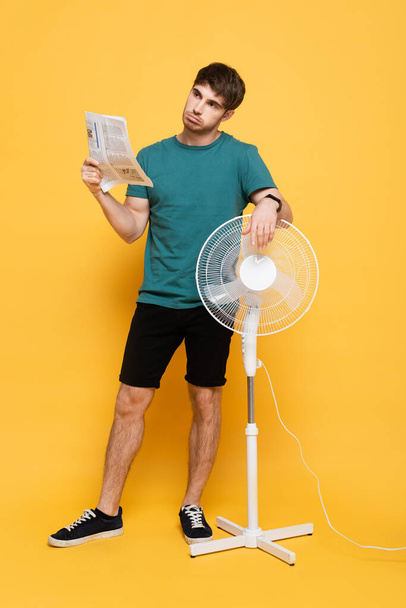 confused man suffering from heat with electric fan and newspaper as hand fan on yellow     - 写真・画像