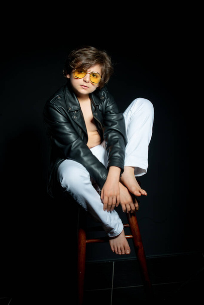 preteen boy in black leather jacket and yellow sunglasses posing on chair on black background - Фото, изображение
