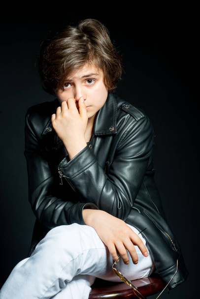 adorable preteen boy in black leather jacket posing on black background and looking at camera - Foto, Bild