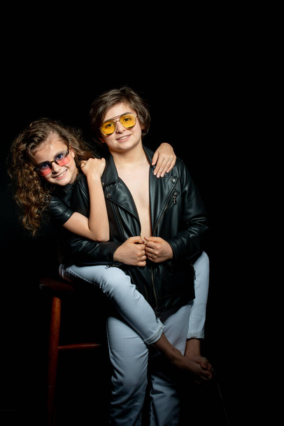 preteen boy and girl in leather clothes and sunglasses posing and hugging in studio on dark background - Photo, Image