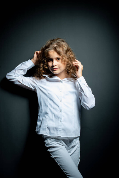 adorable preteen girl in white shirt posing on dark background and looking at camera - Фото, изображение