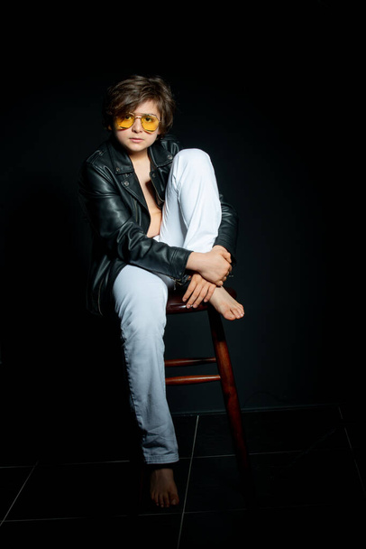preteen boy in black leather jacket and yellow sunglasses posing on chair on black background - Zdjęcie, obraz