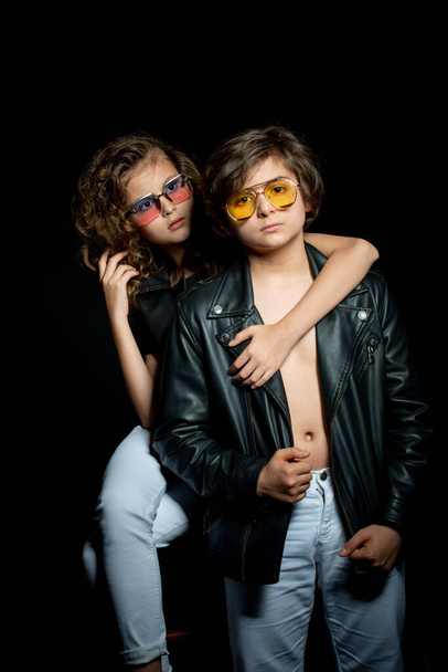 preteen boy and girl in leather clothes and sunglasses posing and hugging in studio on dark background - Photo, image