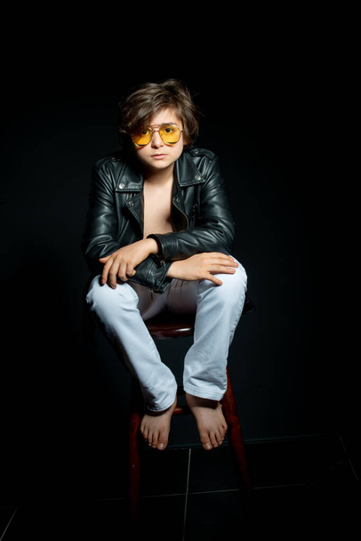 preteen boy in black leather jacket and yellow sunglasses posing on chair on black background - Foto, Imagem