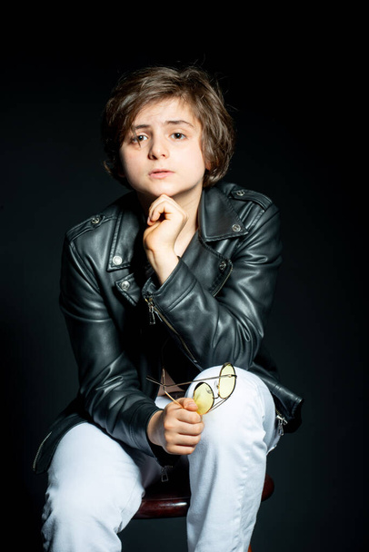 adorable preteen boy in black leather jacket posing on black background and looking at camera - Fotó, kép