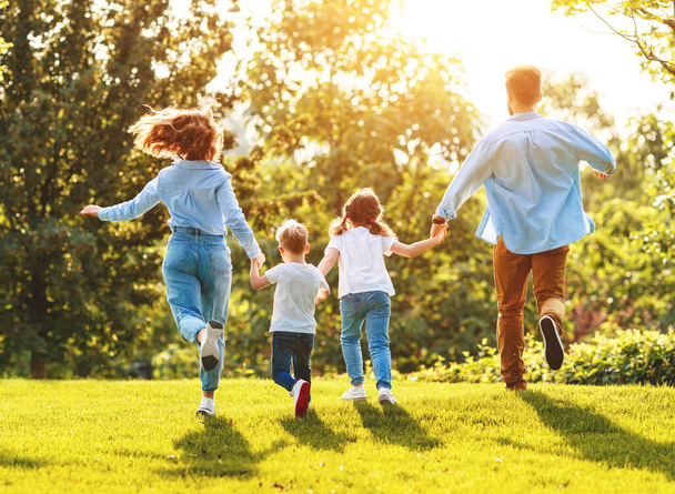 Невідомі щасливі батьки з дітьми в повсякденному одязі бігають назад, тримаючи руки, насолоджуючись разом на зеленій траві влітку
 - Фото, зображення
