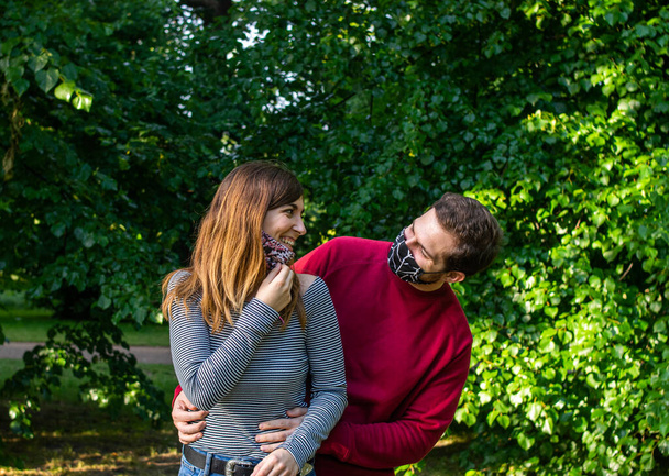 顔のマスクを着て公園でデートをしている若い魅力的なカップルの写真、笑って楽しい時間を過ごして - 写真・画像