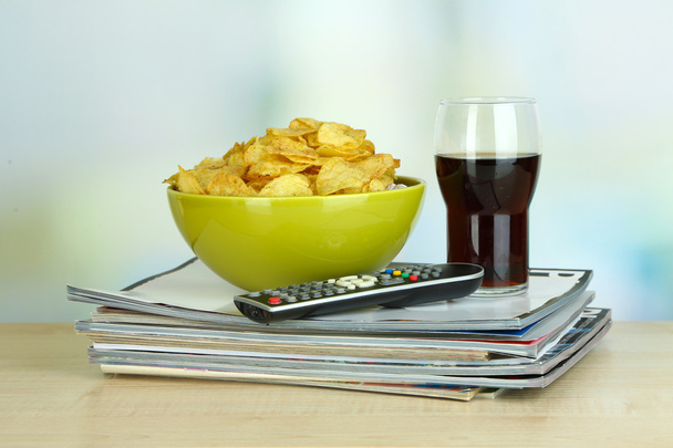 Chips in bowl, cola and TV remote on wooden table on room background - Φωτογραφία, εικόνα
