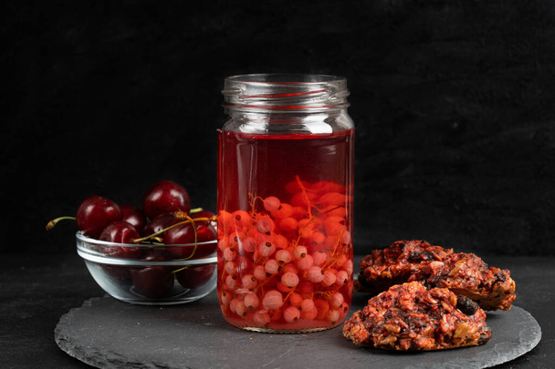 Red currant compote in transparent jar, homemade oatmeal cookies and ripe cherries are laying on stony serving platter against dark background - Photo, image