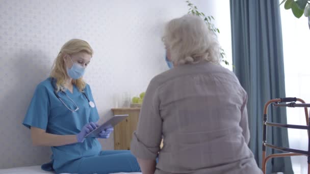 Physician talking to senior patient, putting data in tablet, home consultation - Felvétel, videó