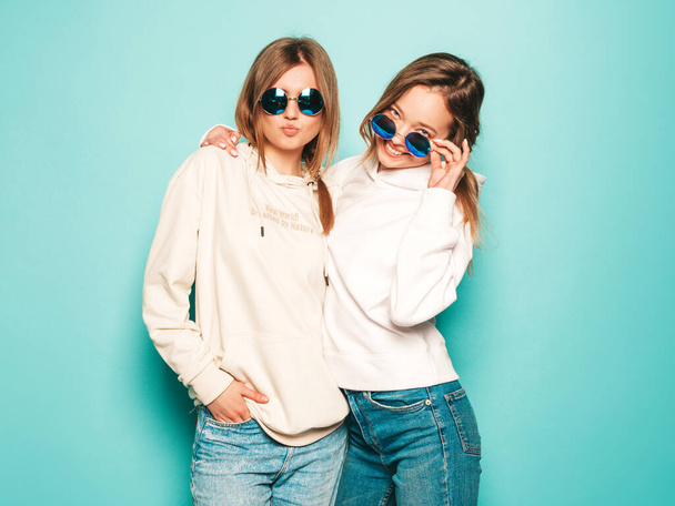 Dvě mladé krásné blond usměvavé hipsterky v módních letních mikinách. Sexy bezstarostné ženy pózující blízko modré zdi. Moderní a pozitivní modely se baví ve slunečních brýlích - Fotografie, Obrázek