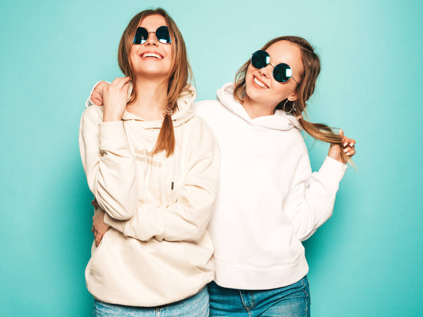 Two young beautiful blond smiling hipster girls in trendy summer hoodie clothes. Sexy carefree women posing near blue wall. Trendy and positive models having fun in sunglasses - Фото, изображение