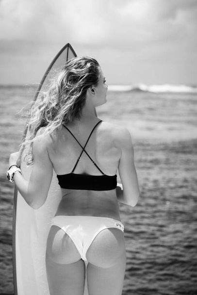 Black and white portrait of young blonde woman in bikini with surfboards on background, she is smiling - Foto, immagini