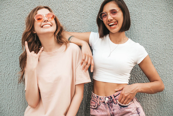 Kaksi nuorta kaunista hymyilevää hipsterityttöä trendikkäissä kesävaatteissa. Seksikkäät huolettomat naiset, jotka poseeraavat katutaustalla aurinkolaseissa. Positiiviset mallit hauskaa ja halaus - Valokuva, kuva
