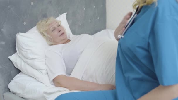 Sick old woman lying in bed talking to young doctor, healthcare services at home - Materiał filmowy, wideo