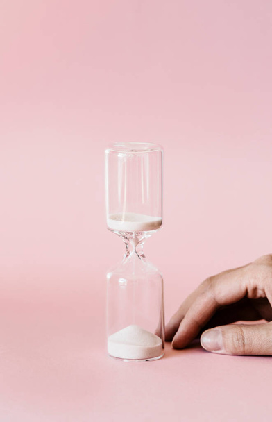 Hourglass and hand on pink background, white sand flowing through the bulb of sandglass measuring time. minimal concept. - Photo, Image