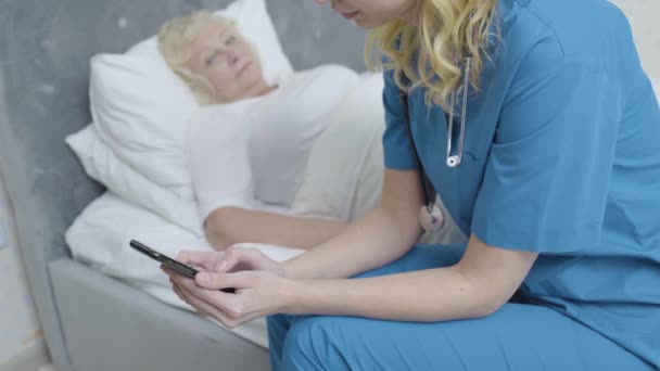 Careless nurse ignoring sick elderly patient, scrolling gadget to waste time - Metraje, vídeo