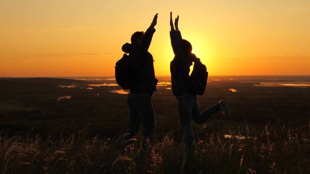 Viajeros, un hombre y una mujer con mochila van al borde de la montaña en rayos del amanecer, levantan las manos en el aire y disfrutan de la victoria, el hermoso sol y el paisaje. Los turistas libres viajan. trabajo en equipo de negocios - Foto, Imagen