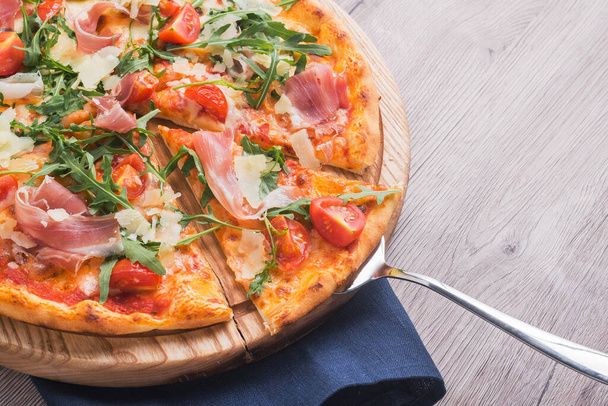 Friss pizza prosciutto hússal, paradicsommal és zöld rugulával egy fa asztali asztalon - Fotó, kép