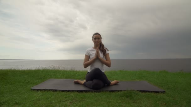 Jeune femme en forme pratique le yoga sur la côte près du lac ou de la mer. Femme assise dans la pose du genou et méditant - Séquence, vidéo