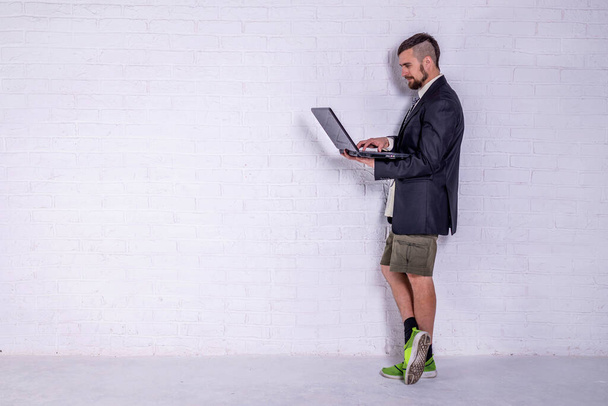 Молодой человек в костюме и шортах работает на ноутбуке, стоя у стены. Концепция удаленной работы. Фрилансер
 - Фото, изображение