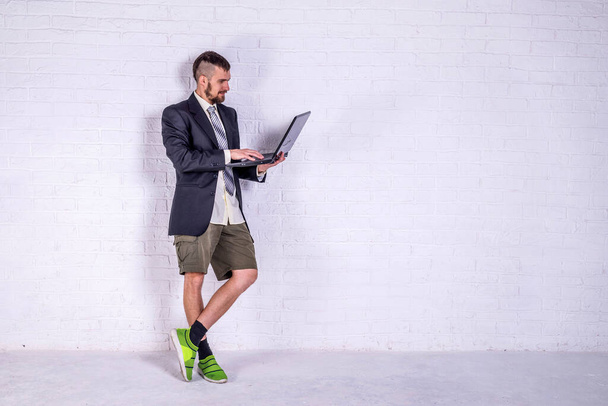 A young man in a suit and shorts works on a laptop while standing near the wall. The concept of remote work. Freelance - Zdjęcie, obraz