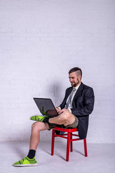 Un joven con traje y pantalones cortos trabaja en un portátil mientras está sentado en una silla cerca de la pared. El concepto de trabajo remoto. Independiente
 - Foto, imagen