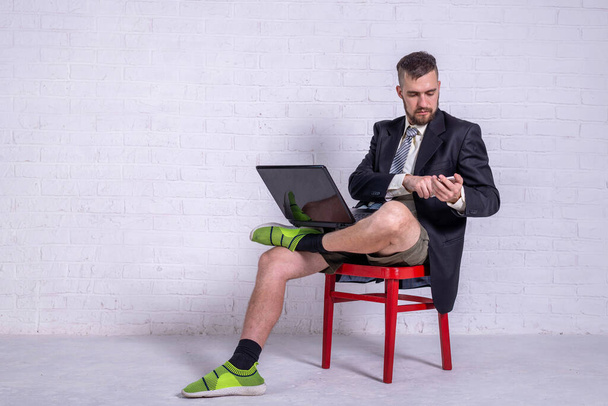 A young man in a suit and shorts works on a laptop while sitting on a chair near the wall, copy space. The concept of remote work. Freelance - Foto, Imagem