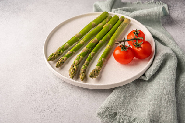 Fresh asparagus and cherry tomatoes on a white plate and napkin on a light backgroun with copy space - Photo, image
