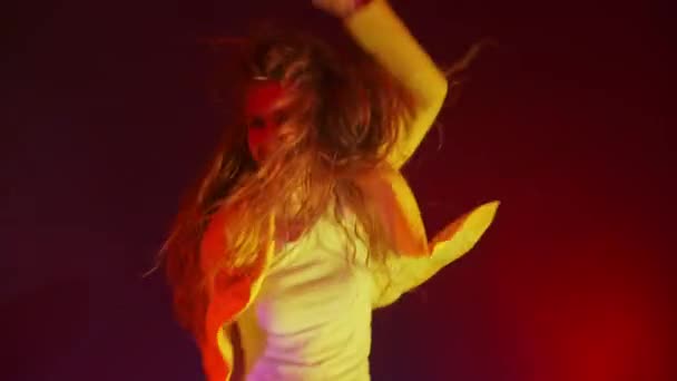 Een grappige vrouw in een geel jasje springt en danst energiek in het neonlicht en rookt - Video