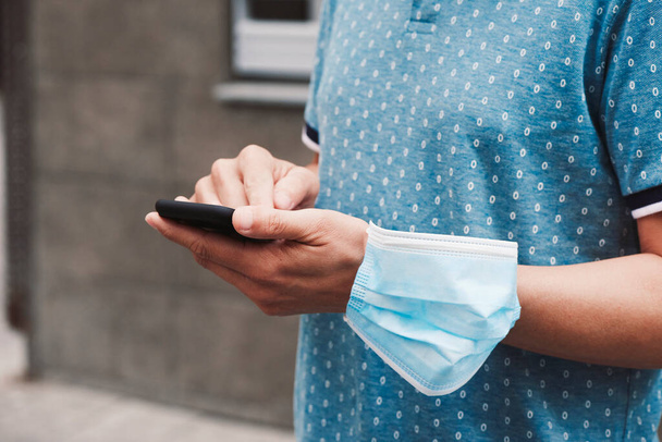 closeup of a young man on the street, in casual wear, using his smartphone and wearing his surgical mask in his arm while is not wearing it in his face - Photo, Image