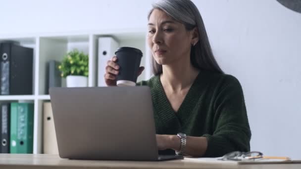 A focused mature woman is using her laptop and drinking coffee while working in the office - Záběry, video