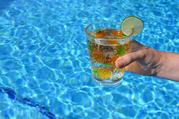 Mujer sosteniendo una taza de plástico decorada con piñas amarillas y una rodaja de lima, en una piscina de color turquesa. vibraciones de verano, mantenerse fresco
. - Foto, Imagen