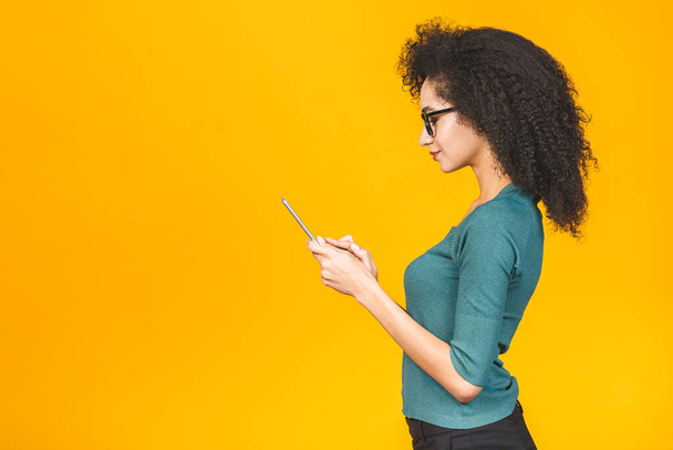 Afrikalı kıvırcık saçlı, dijital tabletli ve gülümseyen genç Amerikalı kız izole edilmiş sarı arka planda duruyor metin, logo ya da reklam için fotokopi alanı var.. - Fotoğraf, Görsel