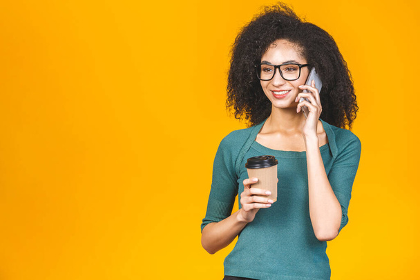 Mutlu Afrikalı genç Amerikalı kadın sarı arka planda tek başına dikilip, cep telefonuyla konuşuyor, kahve ya da çay ikram ediyor.. - Fotoğraf, Görsel