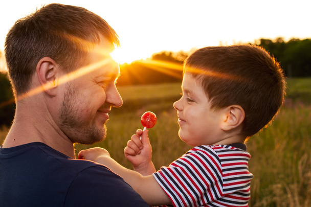 Papa et fils marchent dans un champ d'été jaune le soir au coucher du soleil et mangent des bonbons sur un bâton. Fête des pères, amour dans la famille, le rôle du père dans l'éducation de l'enfant - Photo, image