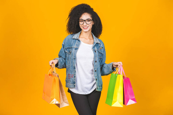 Conceito de compras - Retrato jovem bela mulher afro-americana sorridente e alegre com sacos de compras coloridos isolados sobre amarelo fundo da parede Pastel. Espaço de cópia
. - Foto, Imagem