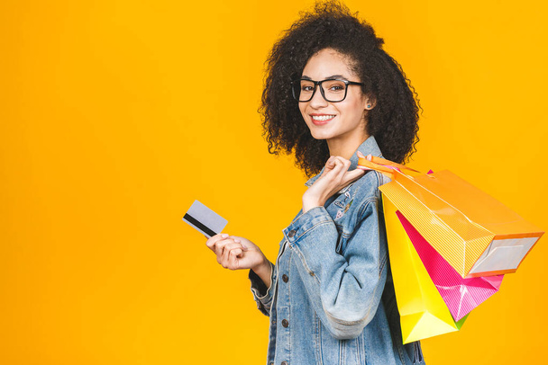 Shopping Concept - Portret jonge mooie Afrikaanse Amerikaanse vrouw glimlachend en vrolijk met kleurrijke boodschappentassen en credit card geïsoleerd over gele achtergrond. Kopieer ruimte.. - Foto, afbeelding