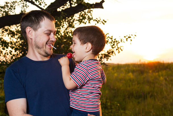 Apa és fia sétálnak egy sárga nyári mezőn esténként naplementekor, és cukorkát esznek egy boton. Apák napja, szeretet a családban, az apa szerepe a gyermek felnevelésében. - Fotó, kép