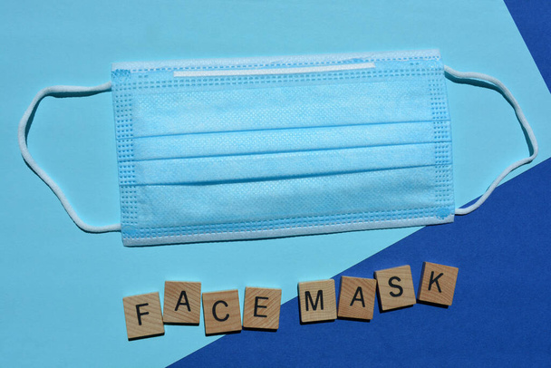Μάσκα προσώπου σε τρισδιάστατα ξύλινα γράμματα αλφαβήτου και μια μη χειρουργική μάσκα προσώπου μιας χρήσης με 3 στρώσεις με ελαστικές θηλιές που απομονώνονται σε μπλε φόντο - Φωτογραφία, εικόνα