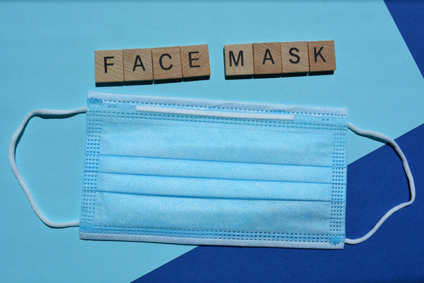 Μάσκα προσώπου, λέξη και μία μη χειρουργική μάσκα προσώπου μιας χρήσης με 3 στρώσεις, με ελαστικές θηλιές αυτιών που απομονώνονται σε μπλε φόντο - Φωτογραφία, εικόνα