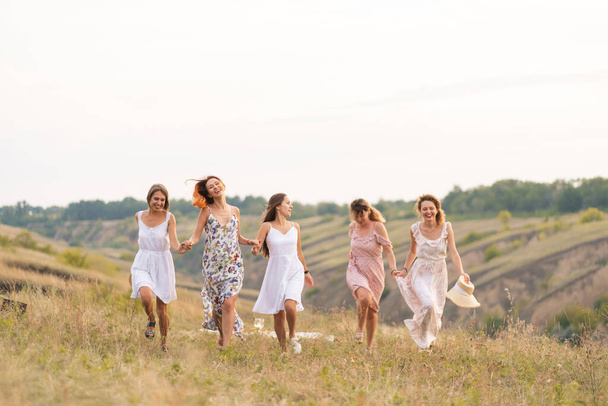 The company of cheerful female friends have a great time together on a picnic in a picturesque place overlooking the green hills. Girls in white dresses dancing in the field. - Fotografie, Obrázek