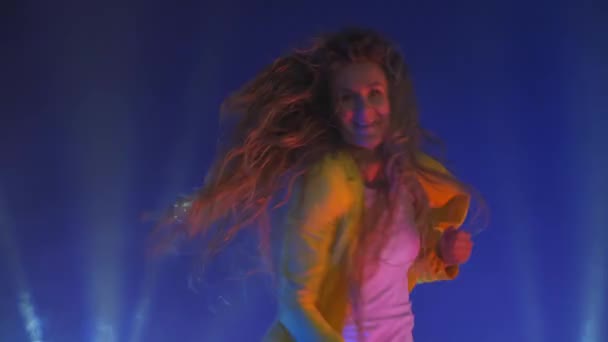 Hauska nainen keltaisessa takissa hyppää ja tanssii energisesti neonin valossa ja savussa. - Materiaali, video