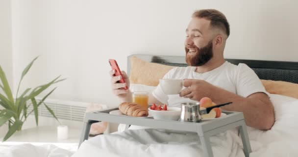 Bearded guy holding coffee cup and reading funny message or joke while lying on bed. Smiling man in 30s using and looking at smartphone screen while having breakfast and laughing. - Imágenes, Vídeo