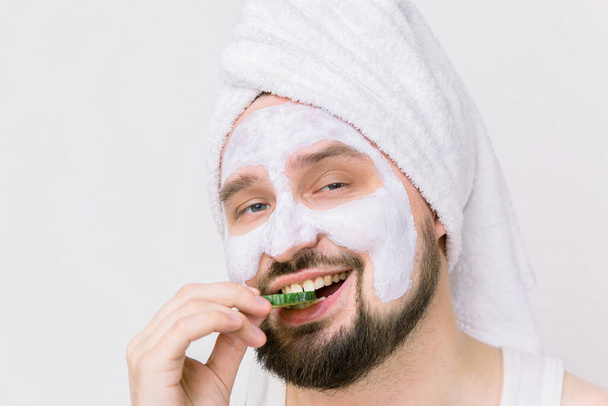 Close up portrait of handsome smiling bearded man in white towel on his head, with mud facial mask after on face, posing with slice of cucumber biting it. Isolated on white background, copy space - Zdjęcie, obraz