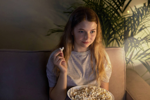 Cute girl sitting on the couch, watching TV with popcorn in a plate. Cozy room. - Fotografie, Obrázek