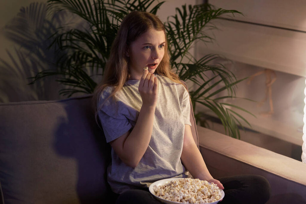 Cute girl sitting on the couch, watching TV with popcorn in a plate. Cozy room. - Fotó, kép