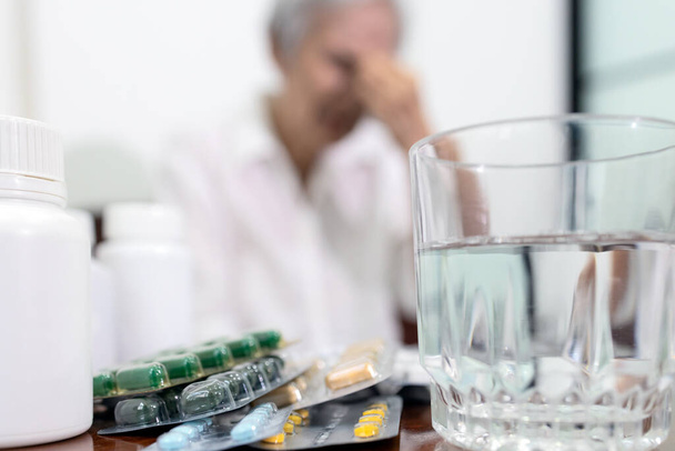 Lot of drugs,medicine bottle,glass of water,treatment medications to reduce pain for senior woman,old elderly in background suffering from migraine,headache with focus on tablets pills or capsules - Photo, Image